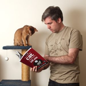 A photo of Theo (an Abyssinian cat) and Matt (a human). Both are looking at a book that's titled "how to tell if your cat is plotting to kill you". Taken in 2013.